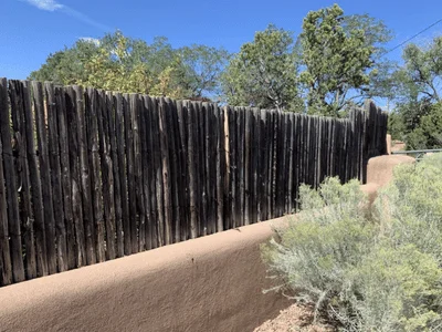 A traditional southwestern coyote fence built with weathered vertical wood stakes sitting on top of a smooth, beige stucco wall.
